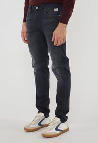 Uomo che indossa jeans slim-fit grigio scuro e sneakers bianche con dettagli blu e beige, in piedi davanti a uno sfondo neutro.