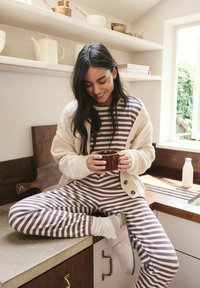 Striped brown and cream pajama set with a fuzzy beige cardigan. Person holds a brown mug, seated on a countertop with wooden accents.