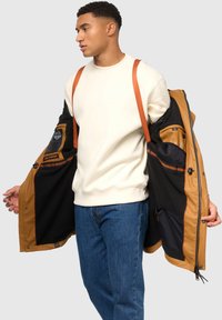 Dunkle Außenjacke mit orangefarbenen Akzenten, gefüttert mit schwarzem Stoff. Das Modell trägt einen cremefarbenen Sweatshirt, blaue Jeans und zeigt das Innere der Jacke.