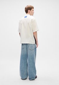 Νέος άνδρας στραμμένος στο πλάι, φορώντας ένα φαρδύ λευκό κοντομάνικο πουκάμισο και φαρδιά ανοιχτόχρωμα μπλε τζιν, σε έναν απλό λευκό φόντο.