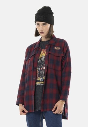 Femme portant un bonnet noir, une veste à carreaux rouge et bleue par-dessus un t-shirt noir graphique, et un jean bleu, debout devant un fond uni.