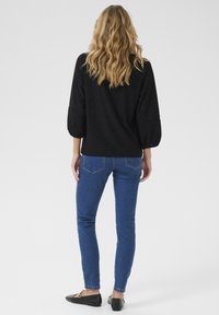 Haut noir texturé à manches bouffantes, associé à un jean skinny bleu et des ballerines noires. Le haut présente des découpes à motifs.