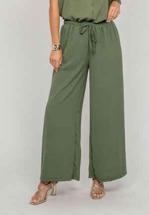 Femme portant un pantalon ample vert olive à taille nouée, un haut assorti, un bracelet en or et des sandales à talons aiguilles à lanières dorées.