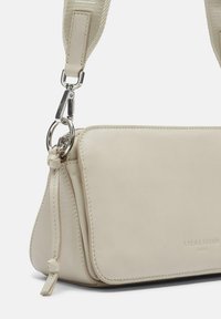 Beige Leder-Crossbody-Tasche mit glatter Textur, dreieckiger Form, silbernem Zubehör und einem gewebten, verstellbaren Tragegurt. Logo des Brands geprägt.