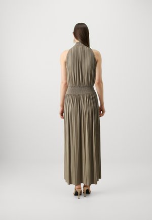 Long, pleated dress in a muted taupe color with a fitted smocked waist and halter neck design. Back view shows flowing skirt and footwear.