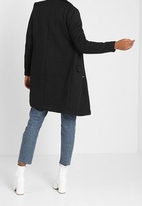 Cappotto lungo nero in tessuto testurizzato, caratterizzato da un taglio dritto, tasca posteriore, abbinato a jeans azzurri chiari e stivali bianchi con tacco alto.