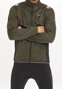 Giacca leggera verde scuro con zip frontale, colletto alto, strisce riflettenti bianche sulle spalle e pannelli laterali neri.