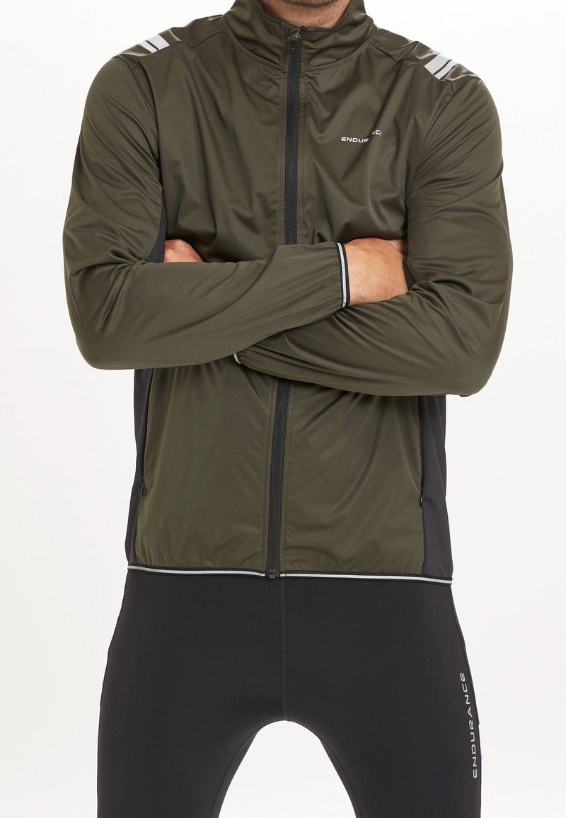 Giacca leggera verde scuro con zip frontale, colletto alto, strisce riflettenti bianche sulle spalle e pannelli laterali neri.
