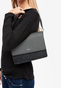 Vrouw in een zwarte longsleeve-top en blauwe jeans, die een grijze en zwarte s.Oliver schoudertas vasthoudt met een getextureerd onderste gedeelte.