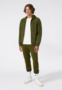 Jongeman die staat te poseren in een olijfgroene ritssluiting hoodie en bijpassende joggingsbroek, een wit T-shirt, witte sokken en witte sneakers op een effen achtergrond.