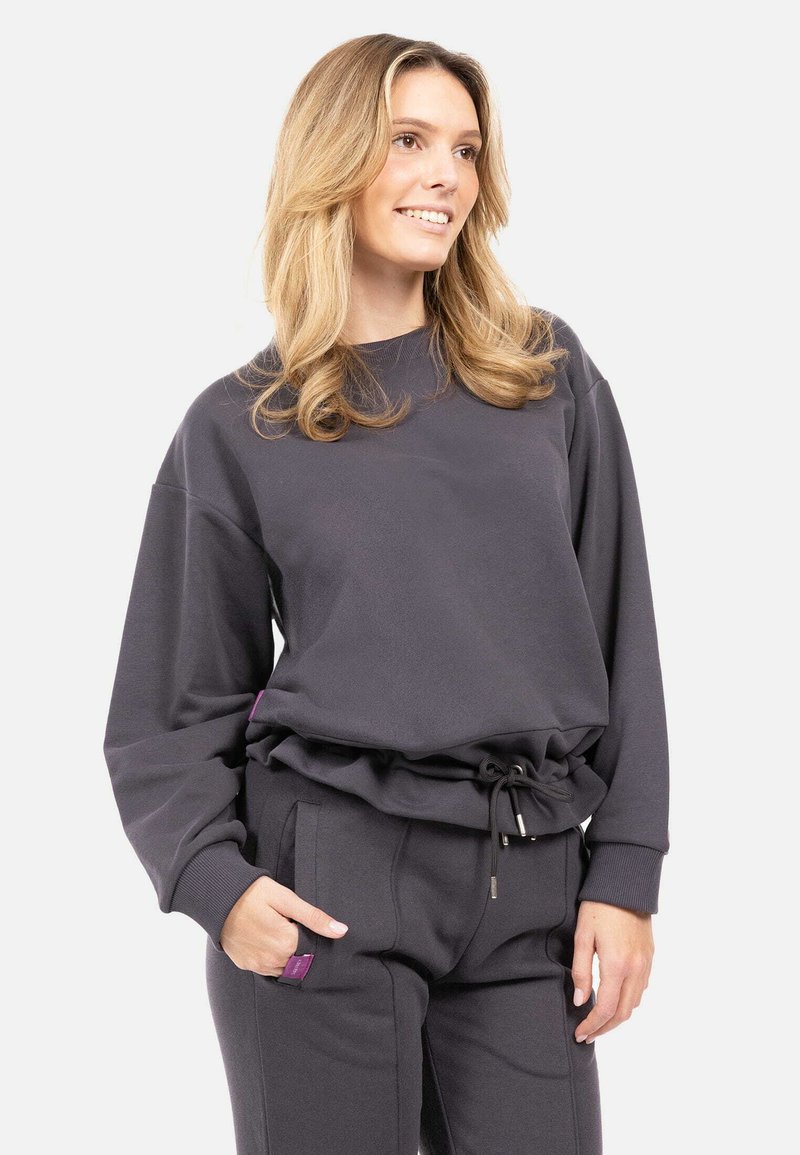 Donna con capelli biondi che sorride, indossando una felpa grigia scura e pantaloni abbinati con vita regulabile, in piedi con una mano in tasca.