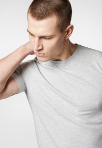 Szara koszulka z krótkim rękawem wykonana z bawełny, posiadająca klasyczny okrągły dekolt i gładką, lekką teksturę. Model pozuje z ręką na szyi.