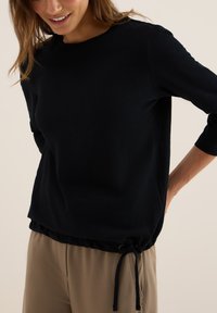 Femme portant un haut noir à manches longues avec un détail de nœud latéral et un pantalon beige, souriante et posant avec une main sur la hanche.