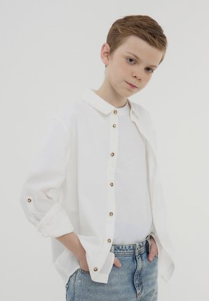 Młody chłopiec o jasnobrązowych włosach, ubrany w białą koszulę, biały t-shirt i jasnoniebieskie dżinsy, z rękami w kieszeniach na jednolitym tle.