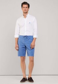 Camicia bianca con maniche arrotolate, pantaloni corti blu con cordoncino e mocassini scuri. Design semplice, tessuto leggero e vestibilità casual.