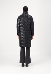 Abrigo largo negro acolchado con textura ondulada, cuello alto y mangas largas, combinado con jeans oscuros acampanados y botas gruesas.