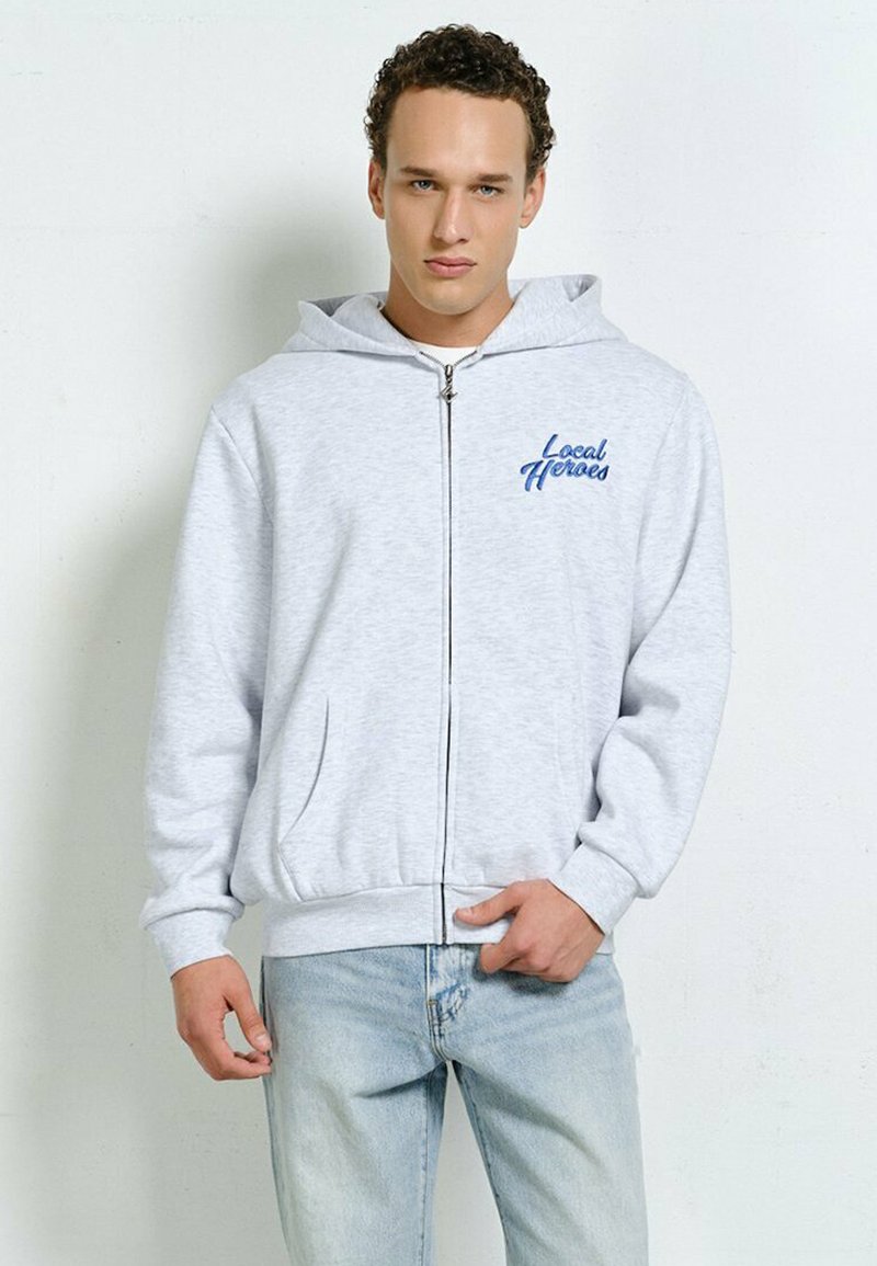 Young man wearing light grey zip-up hoodie with "Local Heroes" text in blue, paired with light blue jeans, standing against plain wall.