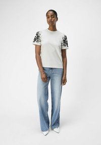 Vit T-shirt med svart blommig broderi på puffärmar, ihop med ljusblå utsvängda jeans och spetsiga vita klackar. Enkelt design, avslappnad passform.