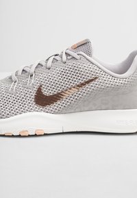 Jasnoszary sportowy but z siateczki o wzorzystej fakturze, brązowymi akcentami, charakterystycznym logo Nike w kształcie haczyka oraz białą amortyzowaną podeszwą.