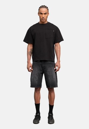 Mann mit geflochtenen Haaren, der ein lockeres schwarzes T-Shirt, ausgewaschene schwarze Jeans-Shorts, schwarze Socken und schwarze Sneakers trägt und vor einem einfarbigen Hintergrund steht.
