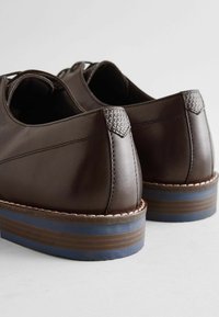 Braune Lederschuhe mit niedrigem Profil, strukturiertem Absatz und kontrastierender blau gestreifter Sohle. Sie verfügen über glatte Oberflächen und detaillierte Nähte.
