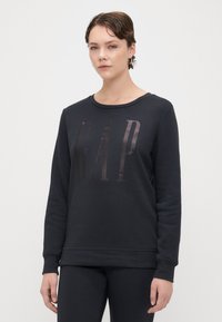 Sudadera negra de suave algodón con cuello redondo, con un logotipo brillante de "GAP" impreso en la parte frontal, mangas largas y puños acanalados.