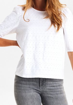 Vrouw met lang haar, gekleed in een wit geperforeerd T-shirt met korte mouwen en grijze spijkerbroek, met één hand rustend op haar rug.