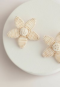 Gold-accented floral earrings with textured white petals and a bead center, displayed on a round, textured white surface.