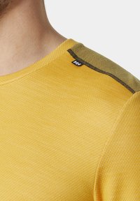 Žluté sportovní tričko s texturovanou tkaninou, kulatým výstřihem a kontrastním detailem na rameni s malou nášivkou.