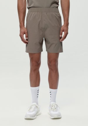 Mand iført taupe sportsshorts med elastisk talje, hvide crew-sokker med mønster og hvide sneakers, stående mod en ensfarvet baggrund.