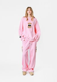 Conjunto de sudadera y pantalones rosa, hecho de una tela suave, con un bordado de oso de peluche en el pecho y bolsillos de carga en los pantalones.
