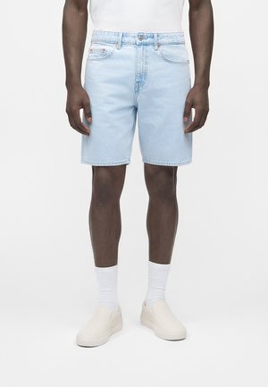 Mężczyzna ubrany w jasnoniebieskie dżinsowe szorty, białe skarpetki, beżowe buty wsuwane i białą koszulę na jednolitym białym tle.