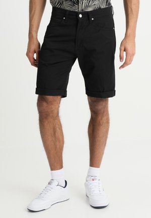 Homme portant un short noir retroussé, des chaussettes blanches et des baskets blanches, debout devant un fond clair uni.