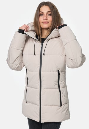 Mujer con una chaqueta acolchada beige con cremallera negra y bolsillos, sujetando la capucha con ambas manos, parada frente a un fondo liso.