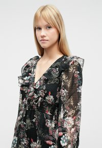 Čierne kvetinové šaty z priesvitnej látky so skladaným výstrihom, dlhými rukávmi a priliehavým pásom s prepracovanými kvetinovými vzormi v rôznych farbách.