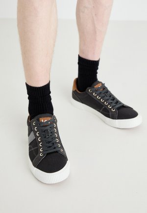 Zapatillas de lona grises con suelas de goma blancas, cordones negros y detalles de cuero marrón en el talón, acompañadas de calcetines negros de canalé.