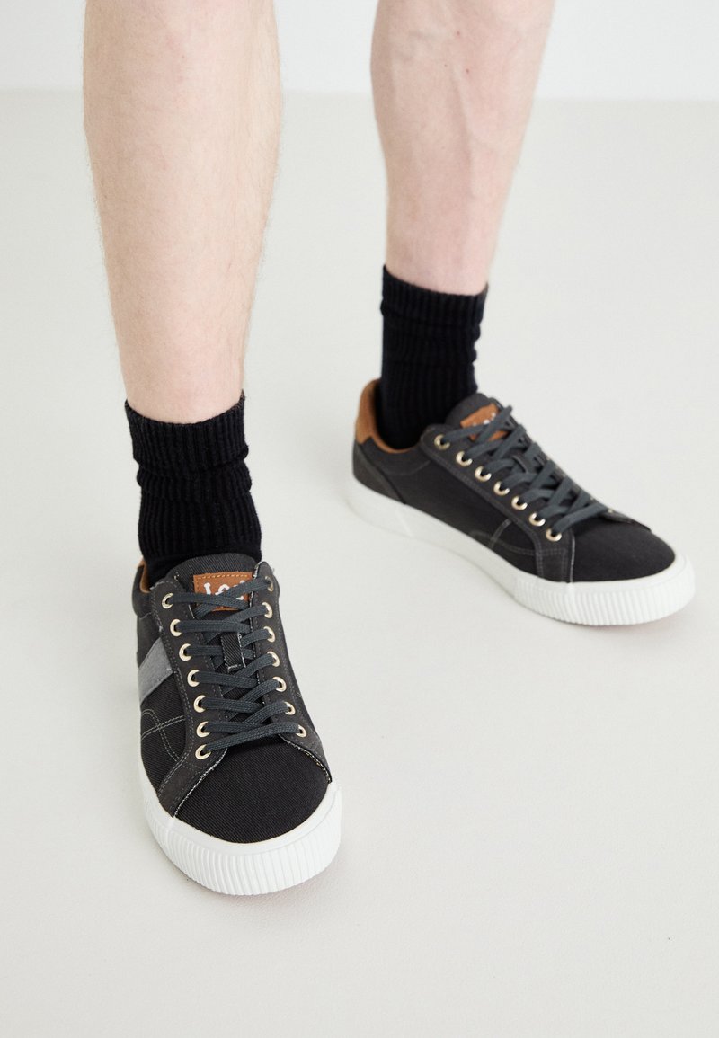 Gray canvas sneakers with white rubber soles, black laces, and brown leather accents on the heel, paired with black ribbed socks.