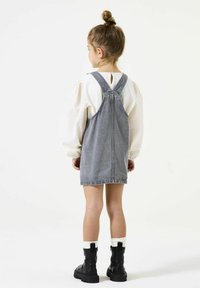 Denim overall jurk, lichtgrijs, A-lijn vorm, verstelbare schouderbanden, rug met sleutelgatdetail. Gekleed met wit bloesje met pofmouwen en zwarte laarzen.