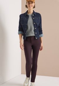 Donkerdenim jack over een grijze geribbelde trui, gecombineerd met bordeauxrode strak zittende broek en witte sneakers, tegen een neutrale achtergrond.