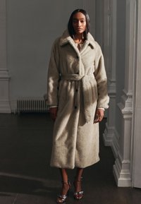 Beige, pluizige jas met een tailleceintuur, grote kraag en zwarte knopen. Zachte textuur en knie-lengte ontwerp. Draagt over een witte top.