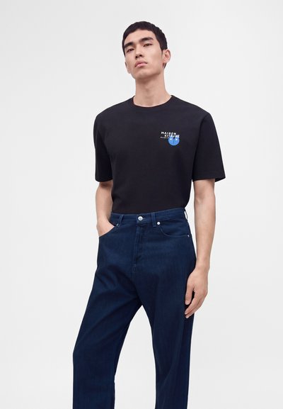 Άνδρας που φοράει μαύρο T-shirt Maison Kitsune και σκούρο μπλε παντελόνι, στέκεται με το ένα χέρι στην τσέπη μπροστά σε απλό φόντο.