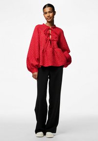 Sarkans auduma blūze ar puffētiem piedurknēm un atslēgas cauruma kakla izgriezumu, pārī ar melniem platām biksēm un baltām sporta kurpēm.