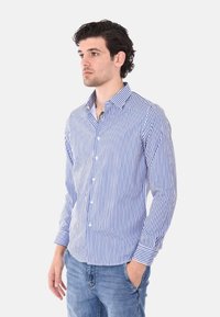 Camicia a righe blu e bianche, con bottoni, maniche lunghe, colletto e orlo arrotondato; abbinata a jeans azzurri chiari.