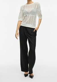 Maglione in bianco con motivo in pizzo intricato e maniche a tre quarti, abbinato a pantaloni neri a gamba larga e scarpe nere a punta.