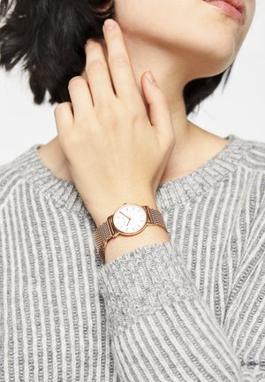 Kvinde rører ved sin nakke med venstre hånd, viser et guldnet armbåndsur og har en grå ribstrikket sweater på.