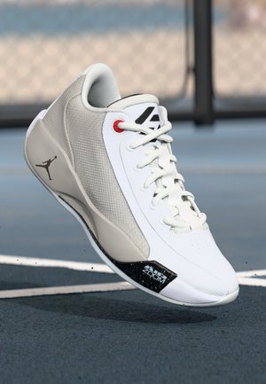 Baskets de sport blanches et grises avec une tige texturée, des accents noirs et rouges, et un logo sur le côté. Elles possèdent un col rembourré et des lacets.