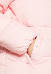 Lichtroze pufferjack met een gestructureerde afwerking. Beschikt over een strakke mouwmanchet en een zijzak met een drukknoopsluiting.