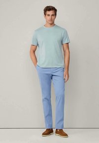 Homem em pé, vestindo uma camisa de manga curta azul clara, calças azul pastel e sapatos castanhos sem atacadores, em fundo plain.