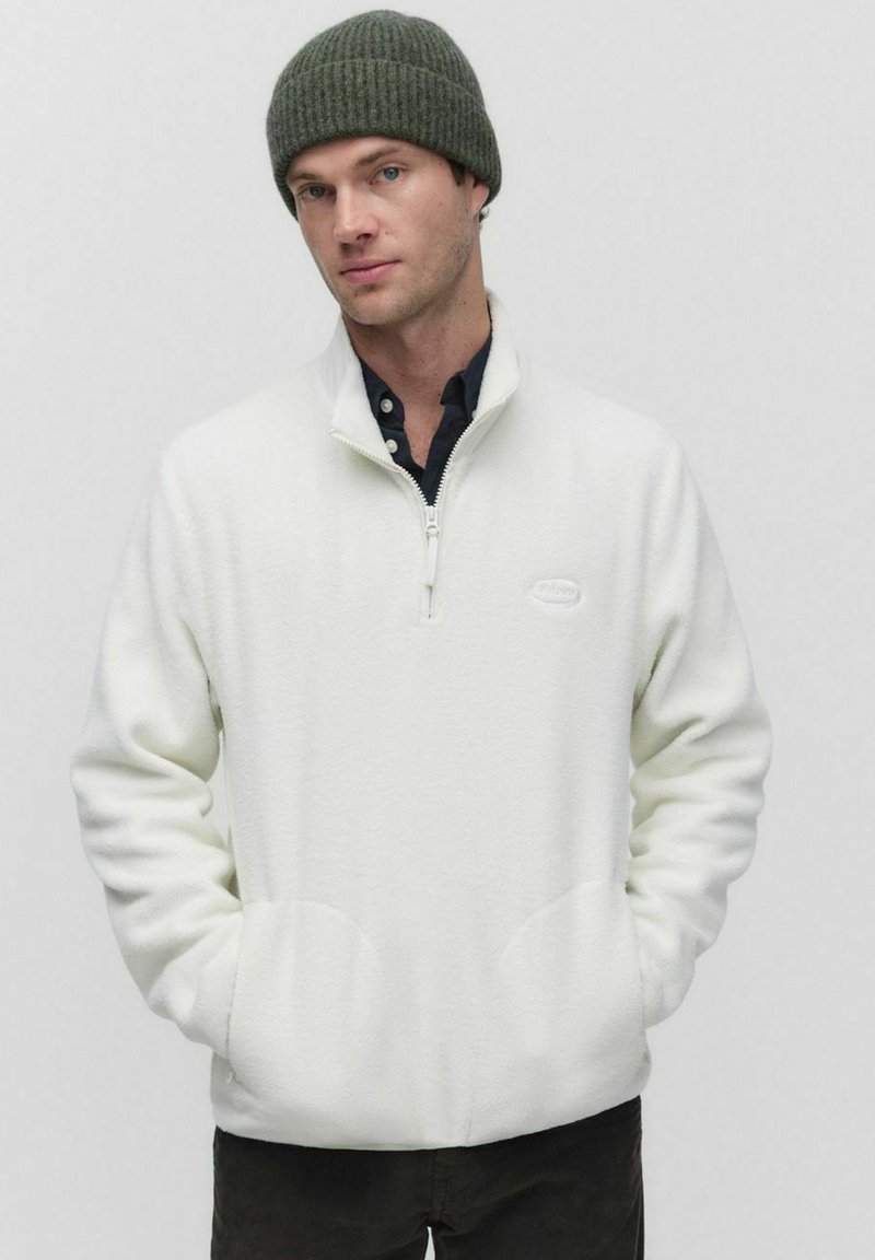 Witte fleece pullover met een kwart-rits, twee voorkiezen en een rond logo. Draagt samen met een donkergroene gebreide beanie. Zachte textuur.