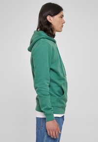 Felpa verde realizzata in tessuto morbido, con tasca frontale, cappuccio con cordoncino e polsini a coste. Indossata con una camicia bianca sotto e jeans blu.
